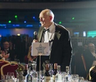 Gerry Weinbren standing with microphone at a table